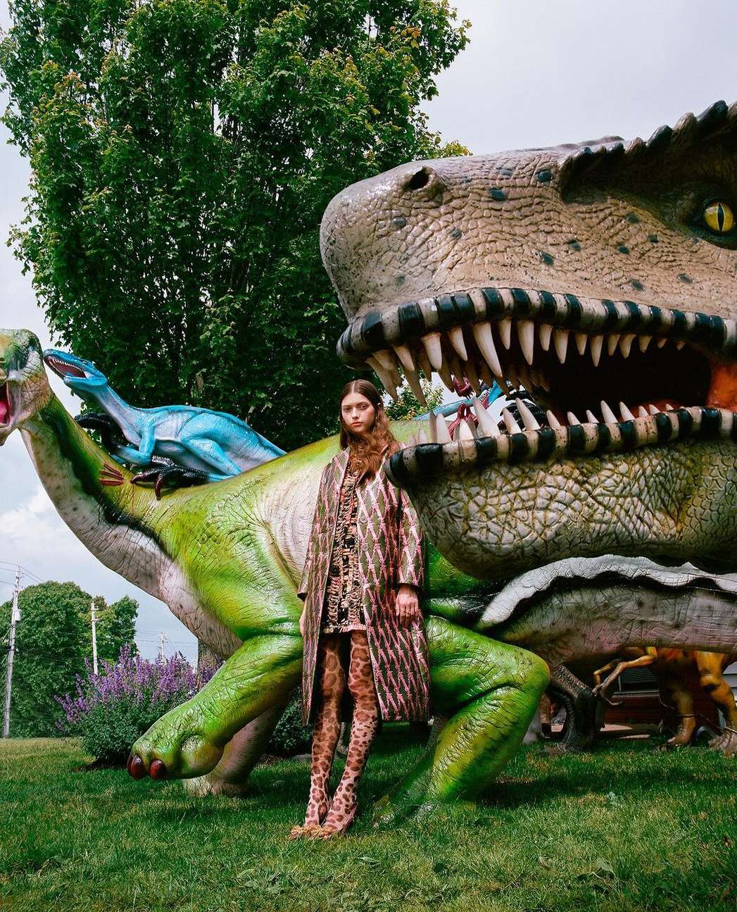 An adult woman in a patterned coat and leggings stands in front of large dinosaur sculptures in an outdoor park-like setting on a cloudy day.
