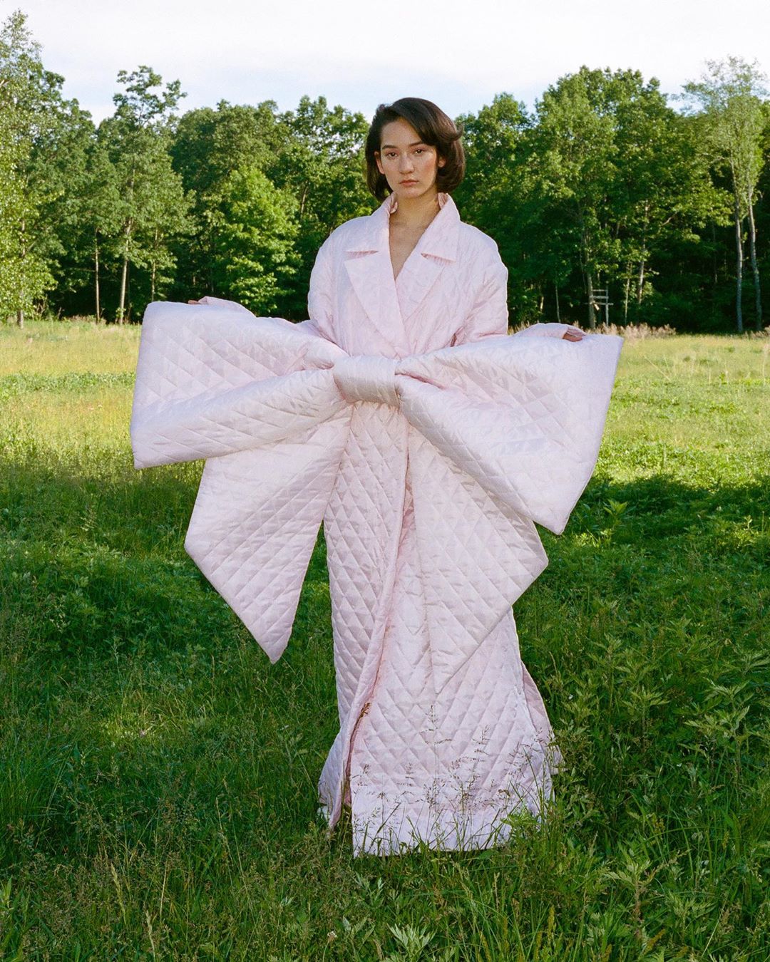 Woman wearing a large pink quilted bow coat standing in a grassy field with trees in the background, outdoor fashion editorial.