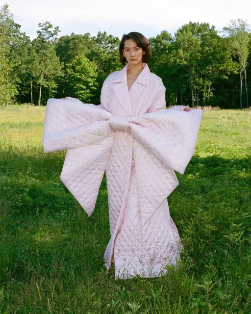 Woman wearing a large pink quilted bow coat standing in a grassy field with trees in the background, outdoor fashion editorial.