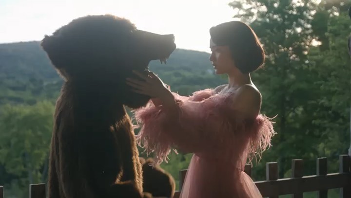 Woman in pink dress interacts with a person in a bear costume outside in natural light with a background of green trees and hills.