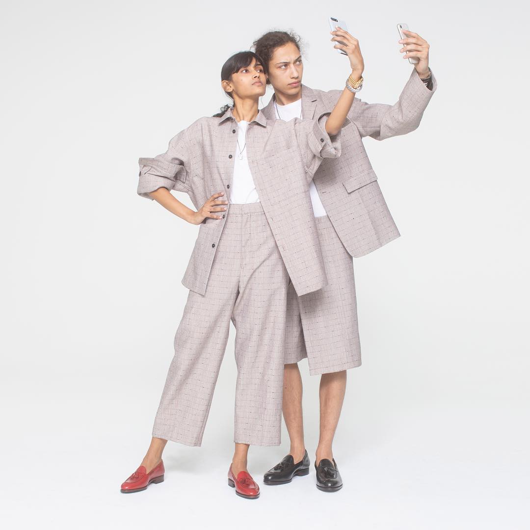 Two people in matching grey plaid outfits take a selfie in a studio, wearing red and black loafers against a plain white backdrop.
