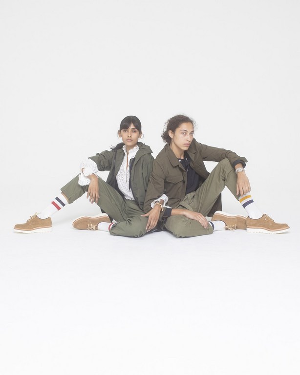 Two adult females in olive green outfits pose seated against a white backdrop, showcasing retro-style fashion with socks and tan shoes.