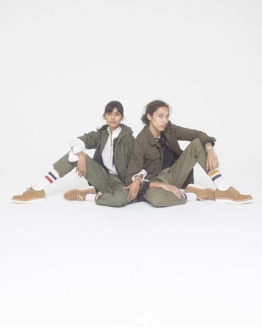 Two adult females in olive green outfits pose seated against a white backdrop, showcasing retro-style fashion with socks and tan shoes.
