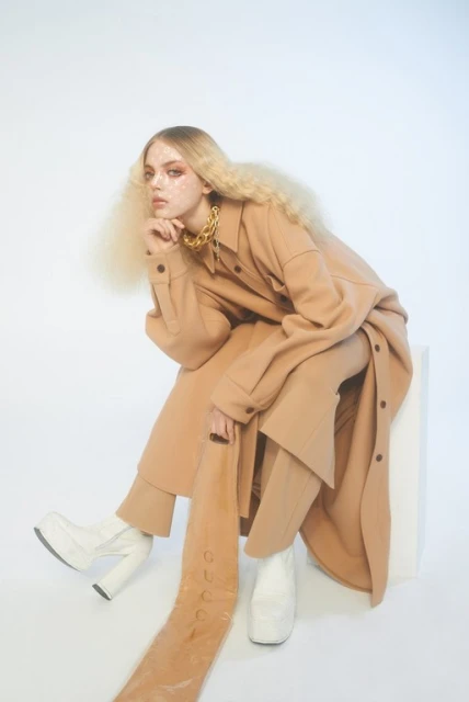 Blonde woman in a tan outfit and white platform boots poses in a photo studio. She wears a chunky gold necklace and sits on a white cube.