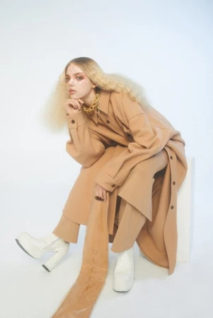 Blonde woman in a tan outfit and white platform boots poses in a photo studio. She wears a chunky gold necklace and sits on a white cube.