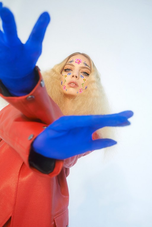 Blonde woman with floral face paint, red jacket, and blue gloves poses against a white background, her hands reaching toward the camera.