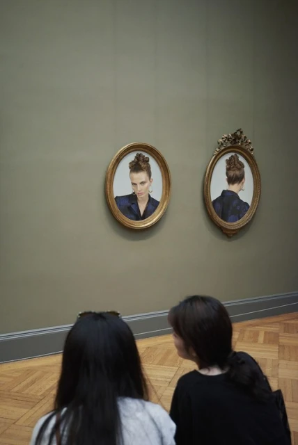 Two women view portrait paintings of a woman with an updo and a suit in an art museum, seen in a vertical shot with neutral colors.