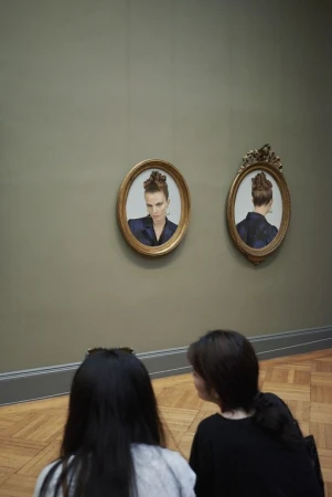 Two women view portrait paintings of a woman with an updo and a suit in an art museum, seen in a vertical shot with neutral colors.