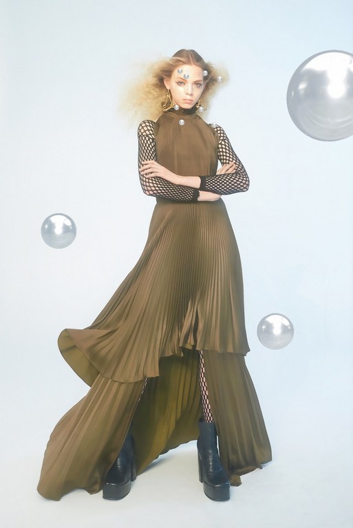 Fashion model poses in a bronze-tone dress with a pleated skirt, black fishnet sleeves, and black platform boots against a pale background.