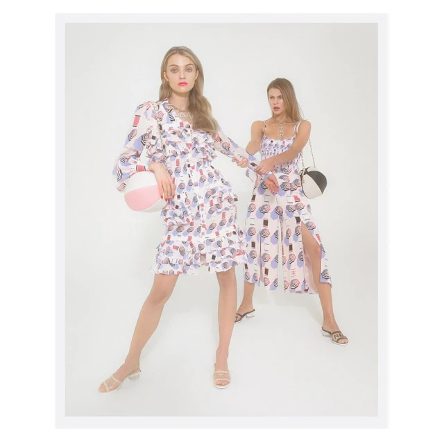 Two women in patterned dresses hold beach balls in a studio setting, showcasing fashion and style with a clean, bright aesthetic.