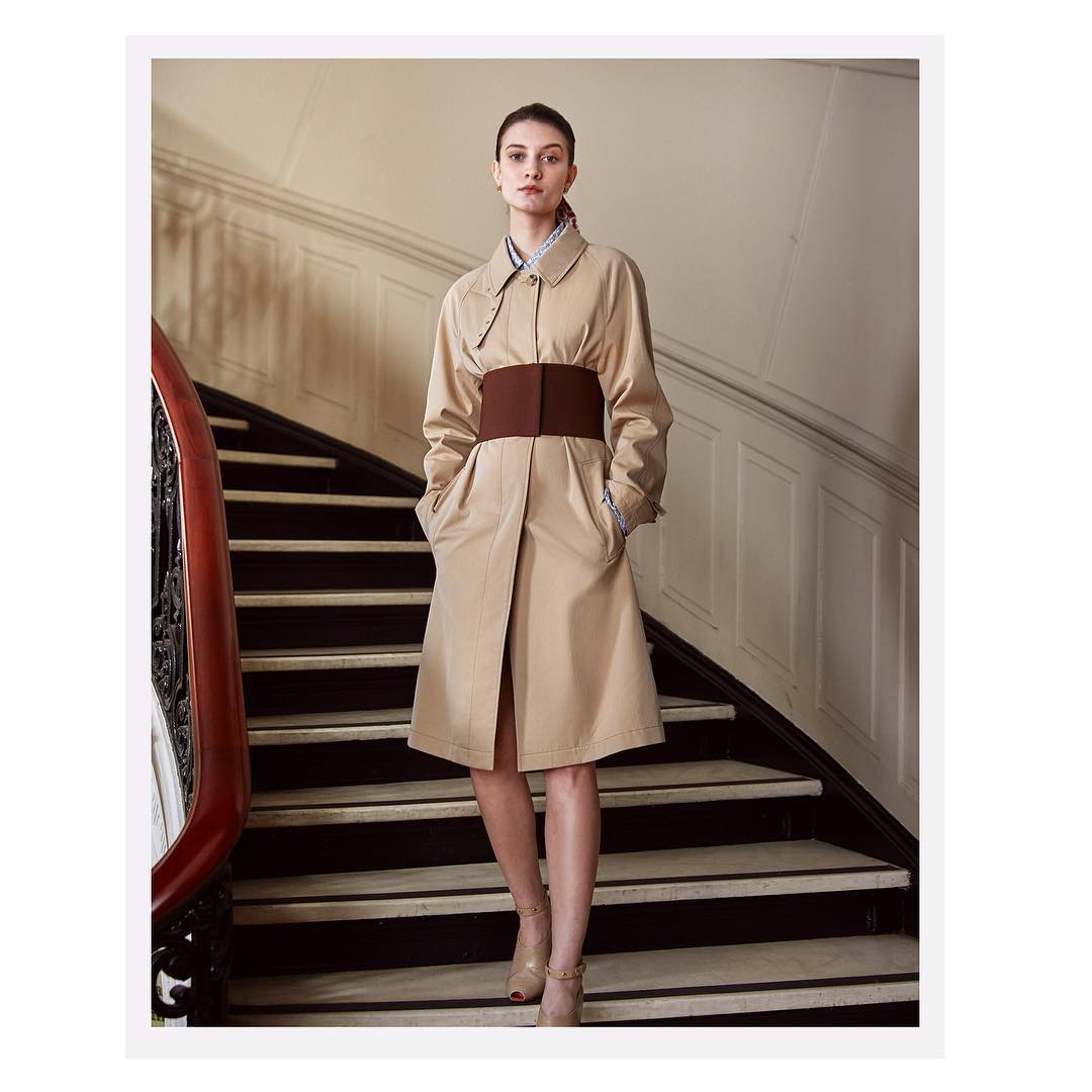 Elegant woman poses on stairs in tan trench coat with brown belt. She stands indoors with hands in pockets and neutral expression.