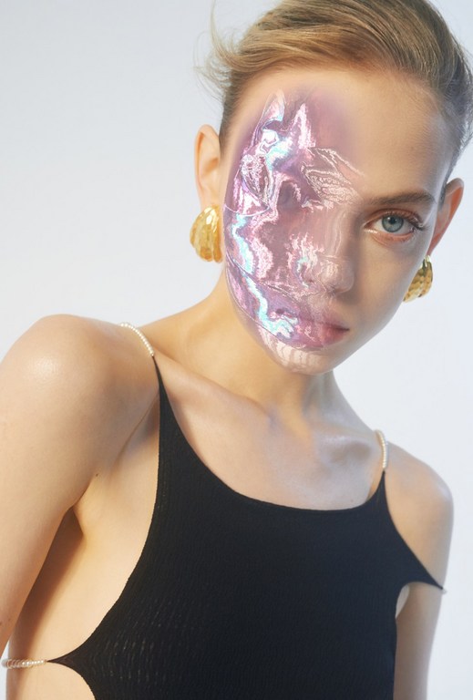 A young fair-skinned woman poses in a studio with an iridescent mask effect over half her face and wearing gold earrings and a black top.