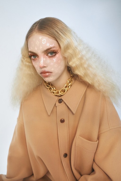 Model with crimped blonde hair wearing a camel shirt and chunky gold necklace, featuring facial monogram print, photographed by Cesar Love Alexandre.