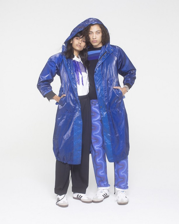Two people wearing blue raincoats and athletic shoes pose in front of a white background in a fashion shot with studio lighting.