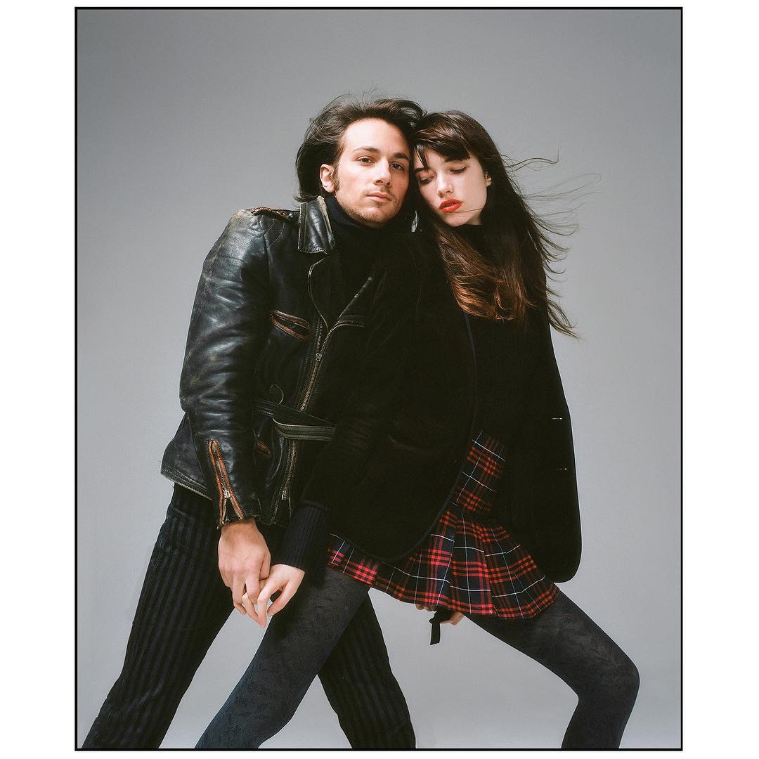 A stylish couple poses indoors against a grey backdrop; the man wears a leather jacket, and the woman wears a blazer and plaid skirt.