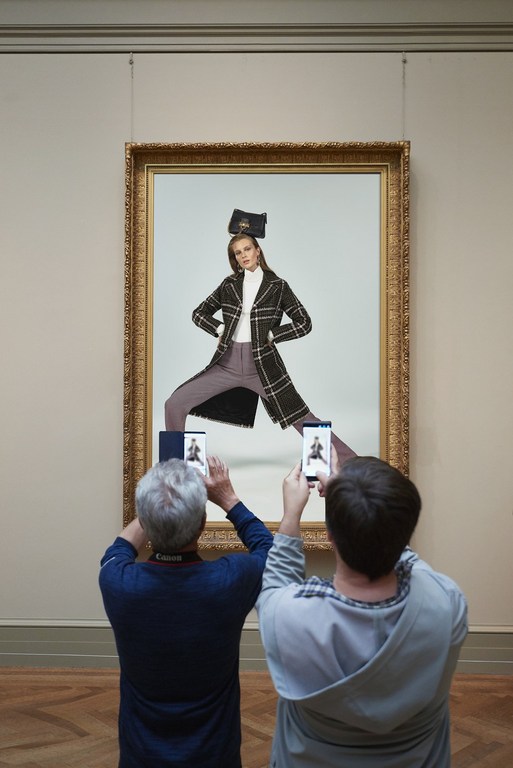 Two people photograph a framed painting of a fashion model in a coat and trousers inside a museum with hardwood floors.