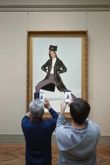 Two people photograph a framed painting of a fashion model in a coat and trousers inside a museum with hardwood floors.