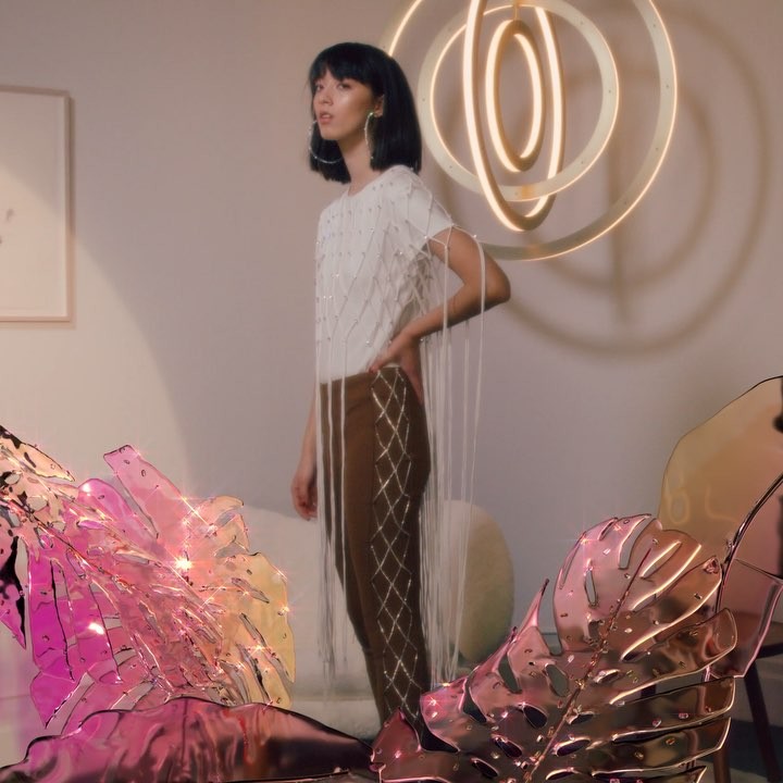 A young adult woman poses indoors wearing a white top with fringe, brown pants, and hoop earrings in a softly lit setting.