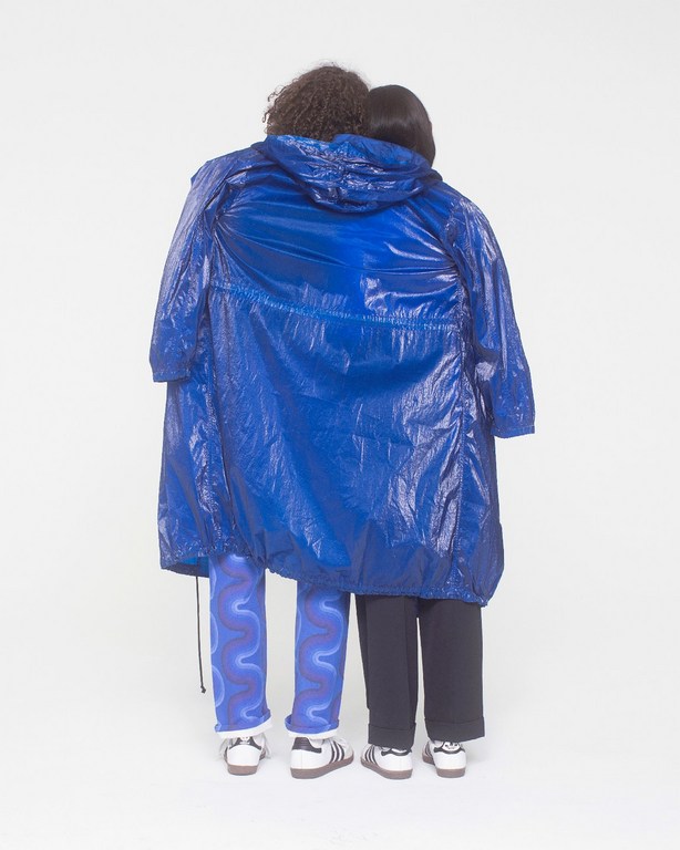 Two individuals stand back-to-back indoors, enveloped in a shiny blue coat, paired with blue patterned and black pants, and sneakers.