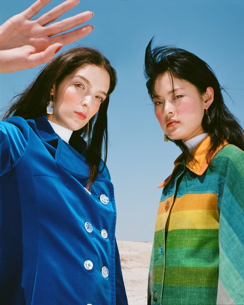 Two women pose outdoors against a light blue sky, wearing colourful jackets. One is wearing a blue velvet jacket, the other rainbow stripes.