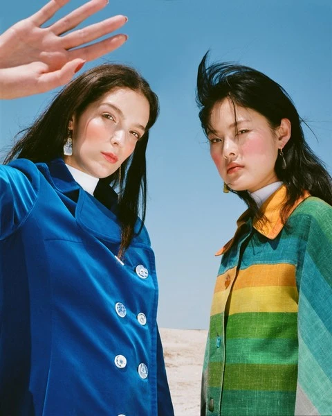 Two women pose outdoors against a light blue sky, wearing colourful jackets. One is wearing a blue velvet jacket, the other rainbow stripes.