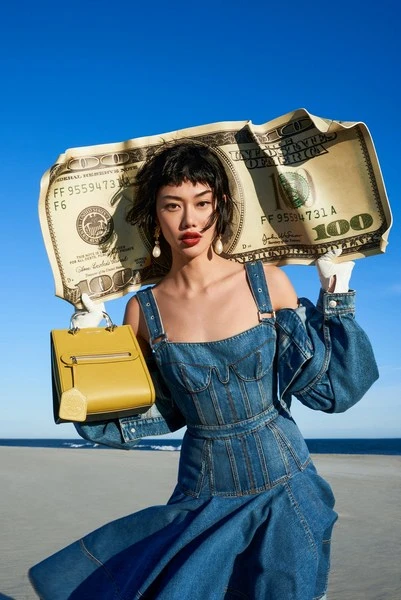Woman with a money prop, wearing a denim dress and holding a yellow purse against a blue sky and neutral sandy beach background.