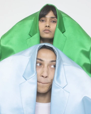 Two models posing with brightly colored blazers pulled over their heads against a plain white background by Cesar Love Alexandre.