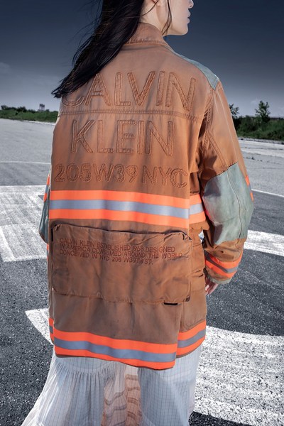 Woman in tan Calvin Klein jacket with orange reflective stripes standing on tarmac with sparse vegetation and overcast sky.