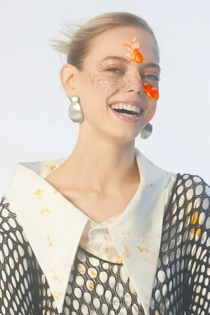 Smiling woman with goldfish graphics on her face, wearing a black mesh top and a large collar. Light and airy, modern aesthetic.