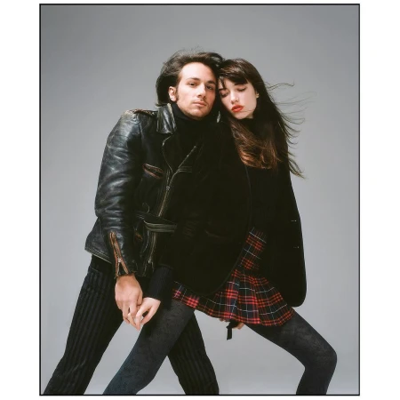 Man in a black leather jacket and woman in a plaid skirt holding hands against a neutral grey background.