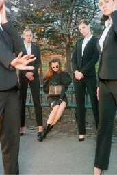 Woman seated on a park bench holding a monogram handbag, surrounded by four standing women in matching black suits.