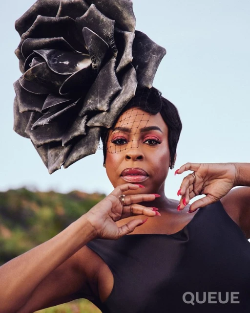 Elegant woman with black floral headpiece poses outdoors with soft natural light, wearing a sleek black dress and subtle makeup.