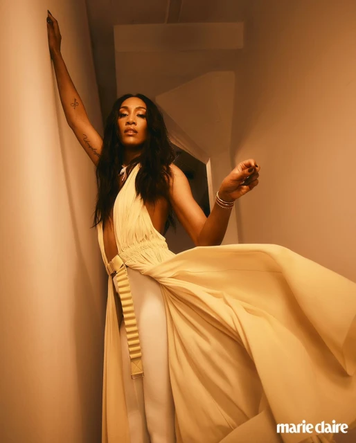 Woman poses in flowing yellow dress with arm raised against neutral wall in indoor setting. Natural lighting, fashion editorial.