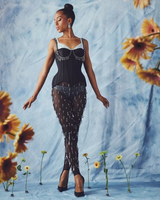 Kerry Washington poses in a black corset, sheer embellished pants, and black heels against a dreamy blue backdrop with flowers.