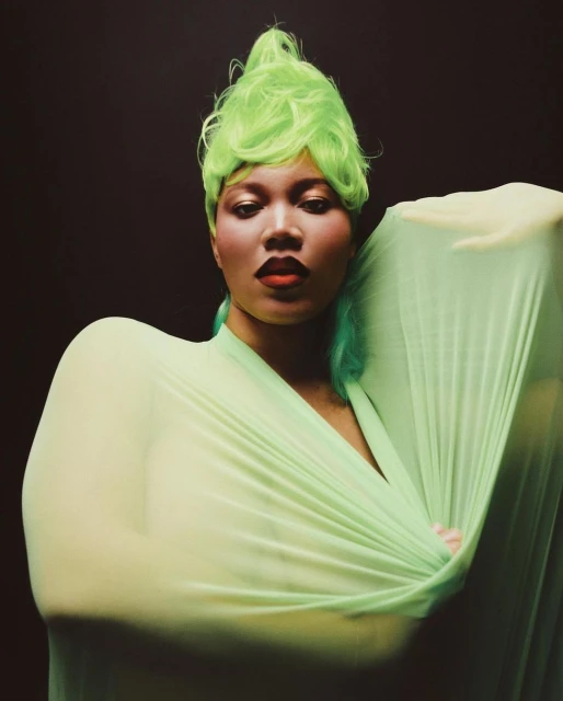 Portrait of a woman with bright green hair and draped in light green fabric against a dark background, showcasing a modern, artistic aesthetic.