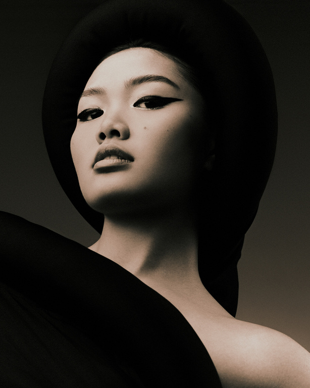 Portrait of a woman with an Asian appearance, wearing a black hat and a draped black garment, set against a dark gray backdrop.