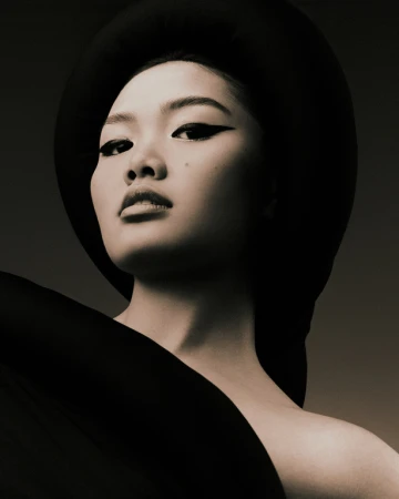 Portrait of a woman with an Asian appearance, wearing a black hat and a draped black garment, set against a dark gray backdrop.