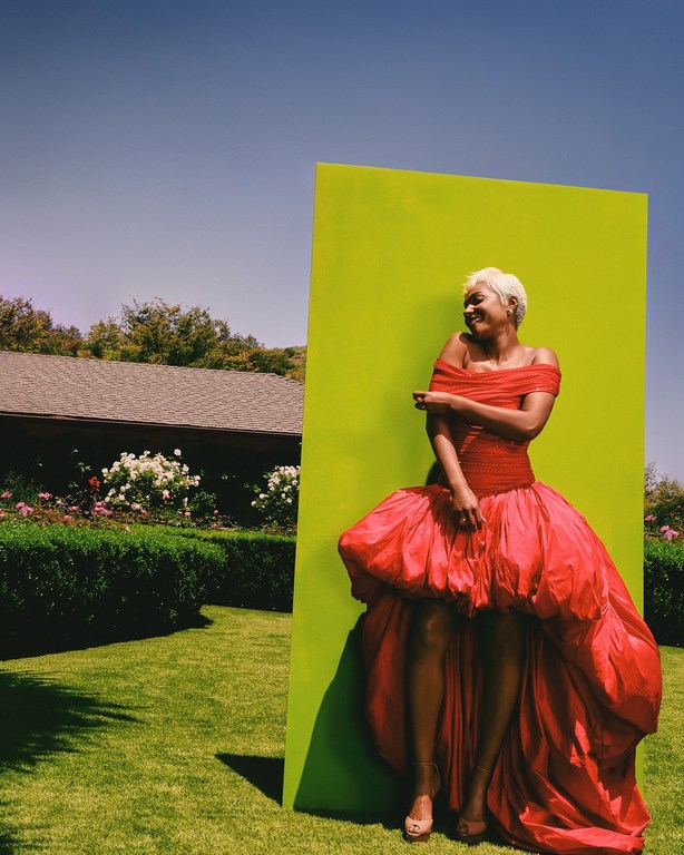 Woman in a red gown poses in front of a lime green backdrop outdoors in a grassy field beneath a blue sky, fashion photography.