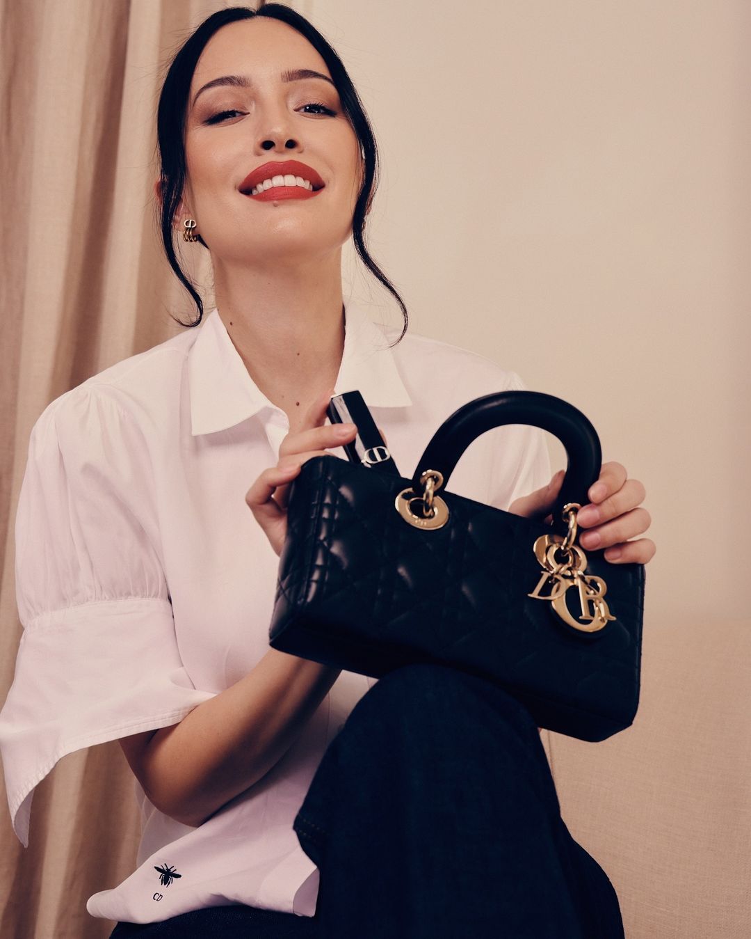 Elegant woman poses with a black leather Lady Dior handbag, wearing a white shirt and black pants in a studio setting.