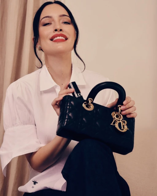 Elegant woman poses with a black leather Lady Dior handbag, wearing a white shirt and black pants in a studio setting.
