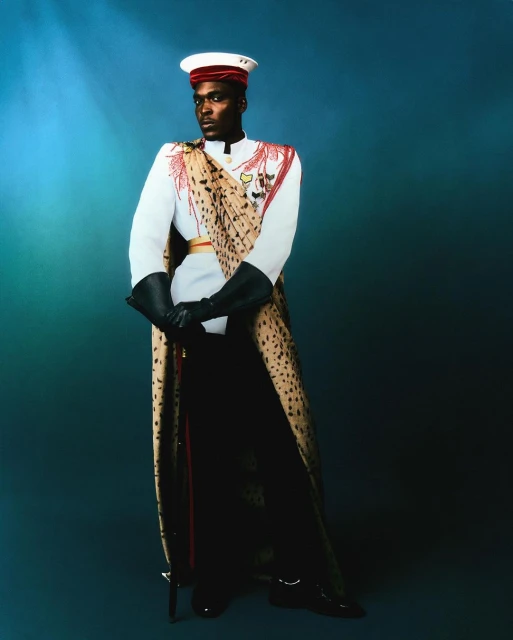 A stately portrait of a Black male model wearing a white uniform with a red hat, gloves, and cheetah-print sash against a teal background.