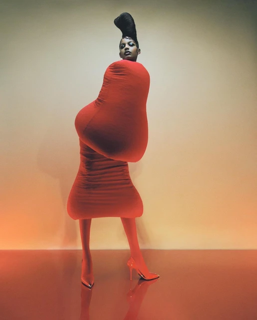 Woman in an avant-garde red fashion ensemble with matching heels poses against a graduated tan backdrop in a studio setting.