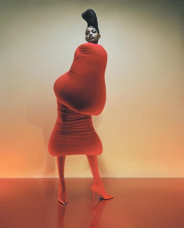 Woman in an avant-garde red fashion ensemble with matching heels poses against a graduated tan backdrop in a studio setting.