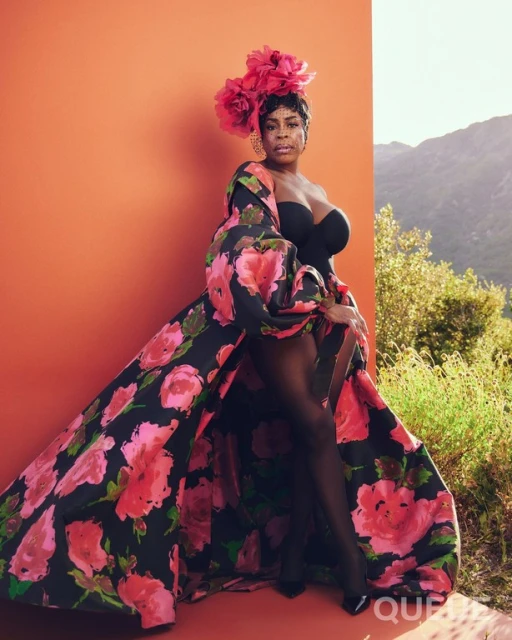 Portrait of an adult woman wearing a floral robe and headpiece, posing in front of a color block of orange and a scenic outdoor view.