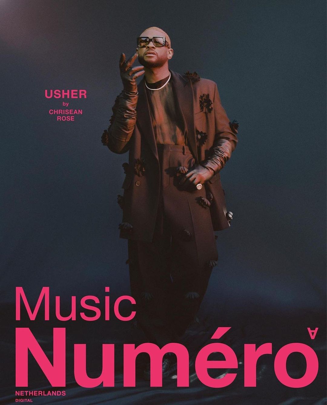 Fashion editorial portrait of a bald adult male, Usher, in a dark jacket with flower accents against a dark teal background. 