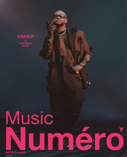 Fashion editorial portrait of a bald adult male, Usher, in a dark jacket with flower accents against a dark teal background. 