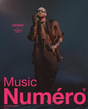 Fashion editorial portrait of a bald adult male, Usher, in a dark jacket with flower accents against a dark teal background. 