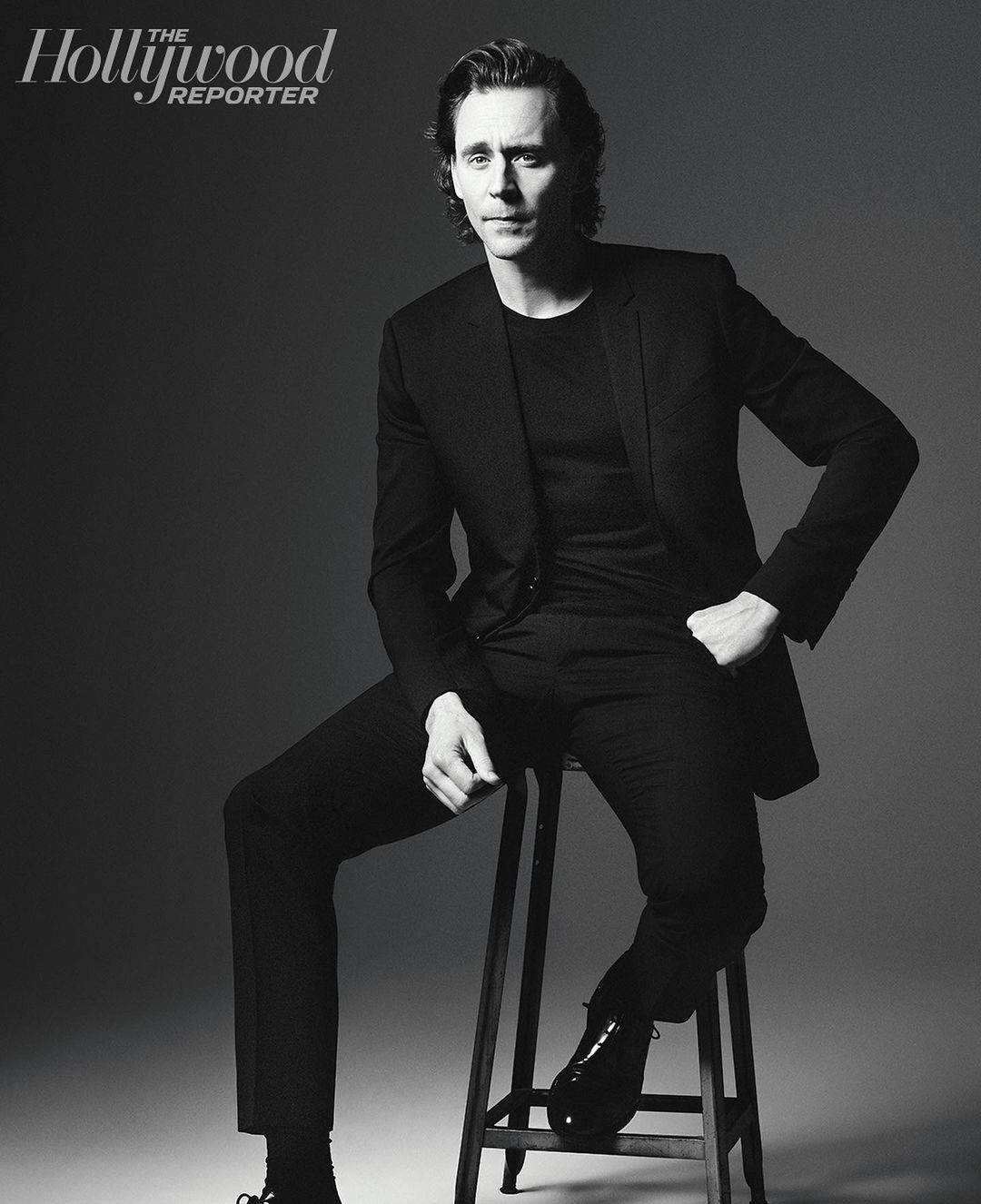 Portrait of Tom Hiddleston in black suit, seated on stool against gray backdrop, Hollywood Reporter logo visible. Studio lighting.