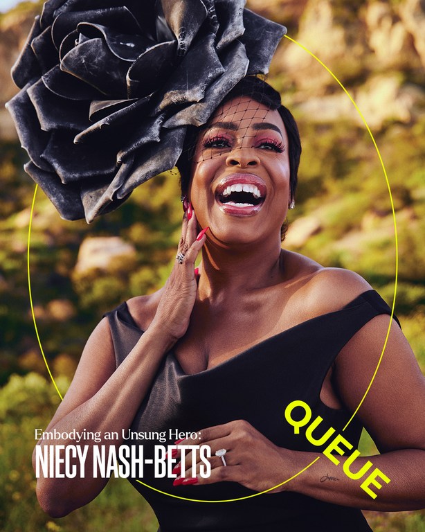 Niecy Nash-Betts smiles in a medium shot outdoors, wearing a black dress and a large flower headpiece with a veil. 