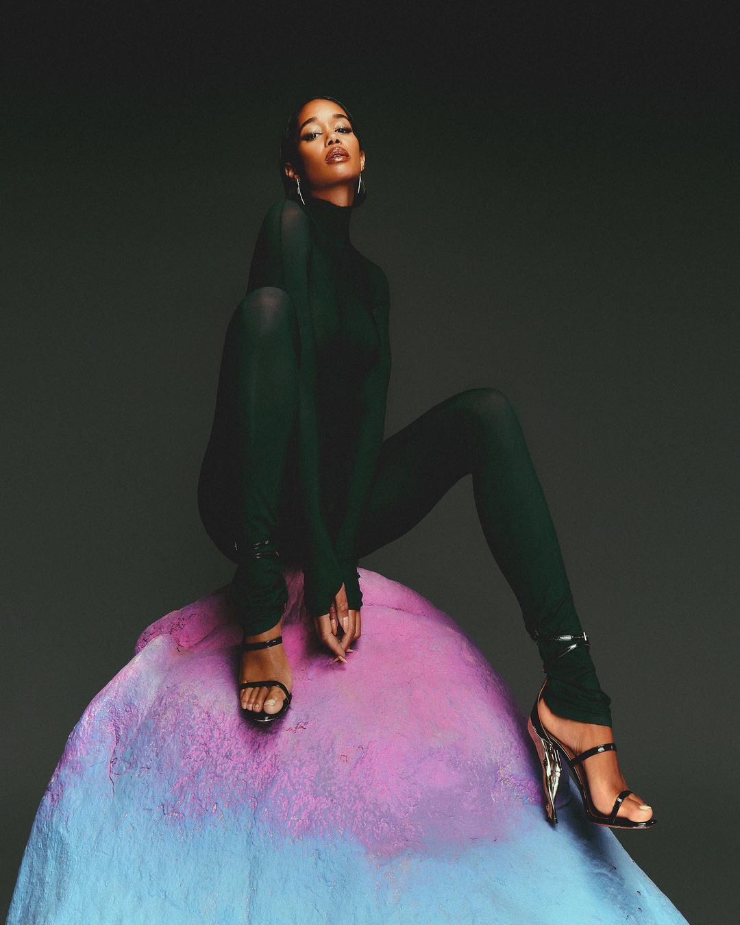 Adult female in stylish green bodysuit and high-heel sandals sits on a colorful stone-like form against a dark gray studio backdrop.